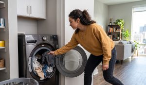 Woman in a mustard sweater loading clothes into a front-loading washer in a bright apartment laundry alcove.